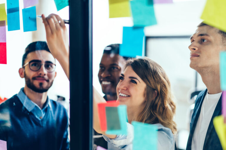 Smiling cheerful female entrepreneur writing funny information on paper stick while enjoying cooperation with male entrepreneur colleagues, multicultural people creating new strategy for proud ceo